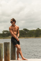 A shirtless man wearing black swim trunks and sunglasses stands barefoot on a dock by the water, with a cloudy sky and tree line in the background.