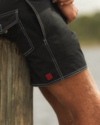 Close-up of a person wearing black board shorts with white contrast stitching, leaning against a wooden dock post near water.