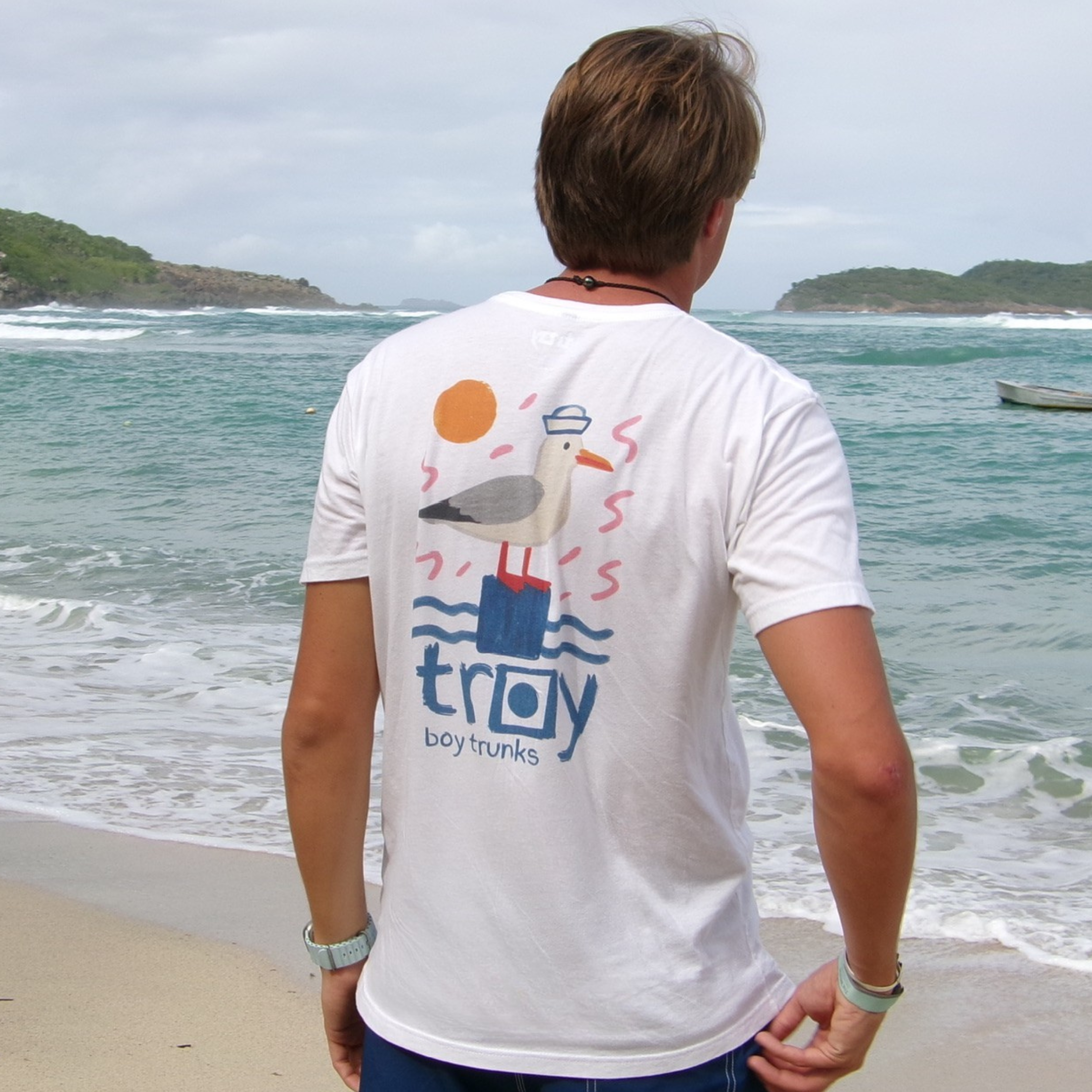 Person wearing a white t-shirt with colorful graphics and 'troy' branding on a beach.